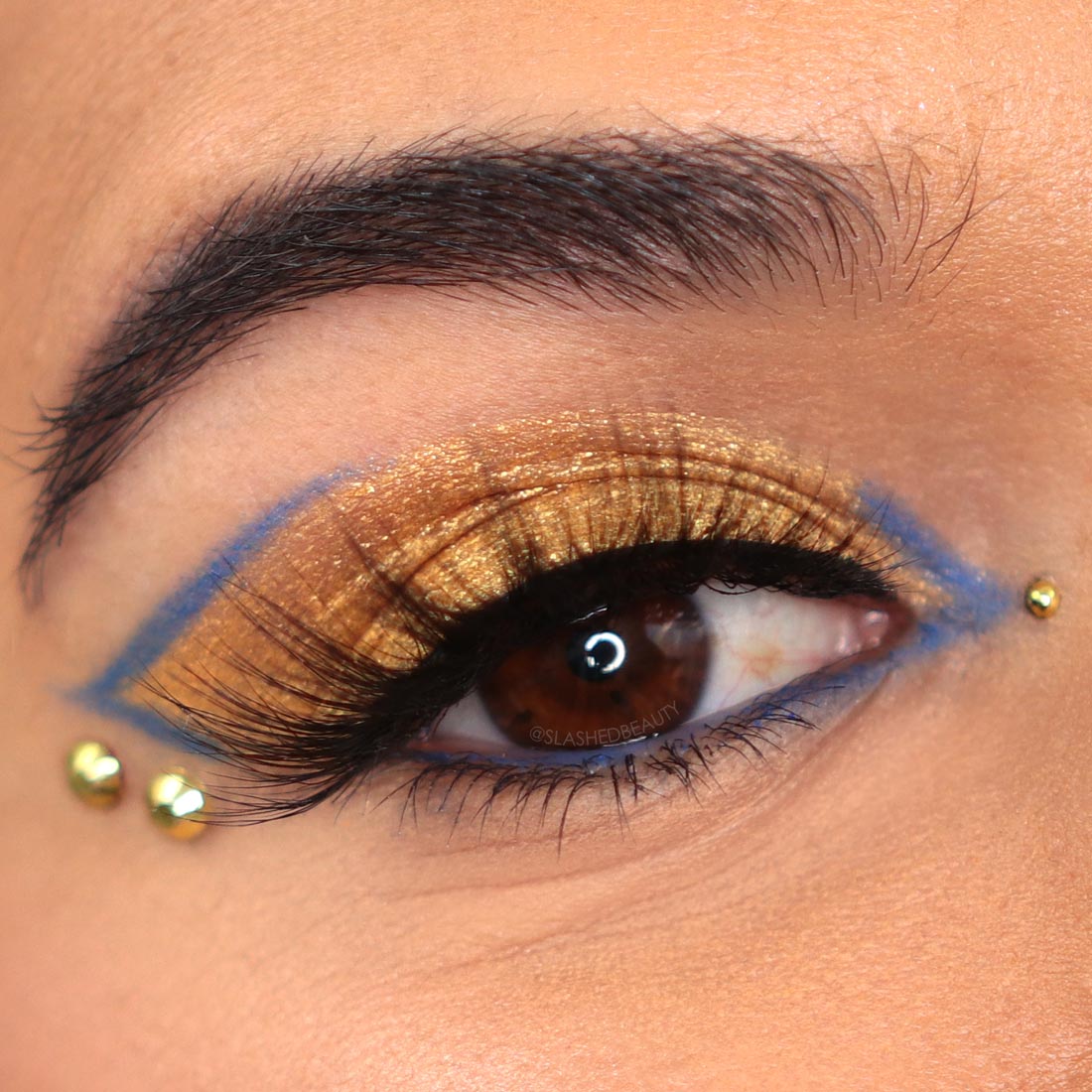 Close up of eye wearing a gold and blue graphic eyeliner look and eye gems | 5 Daring 2022 Beauty Trends to Try This Year | Slashed Beauty
