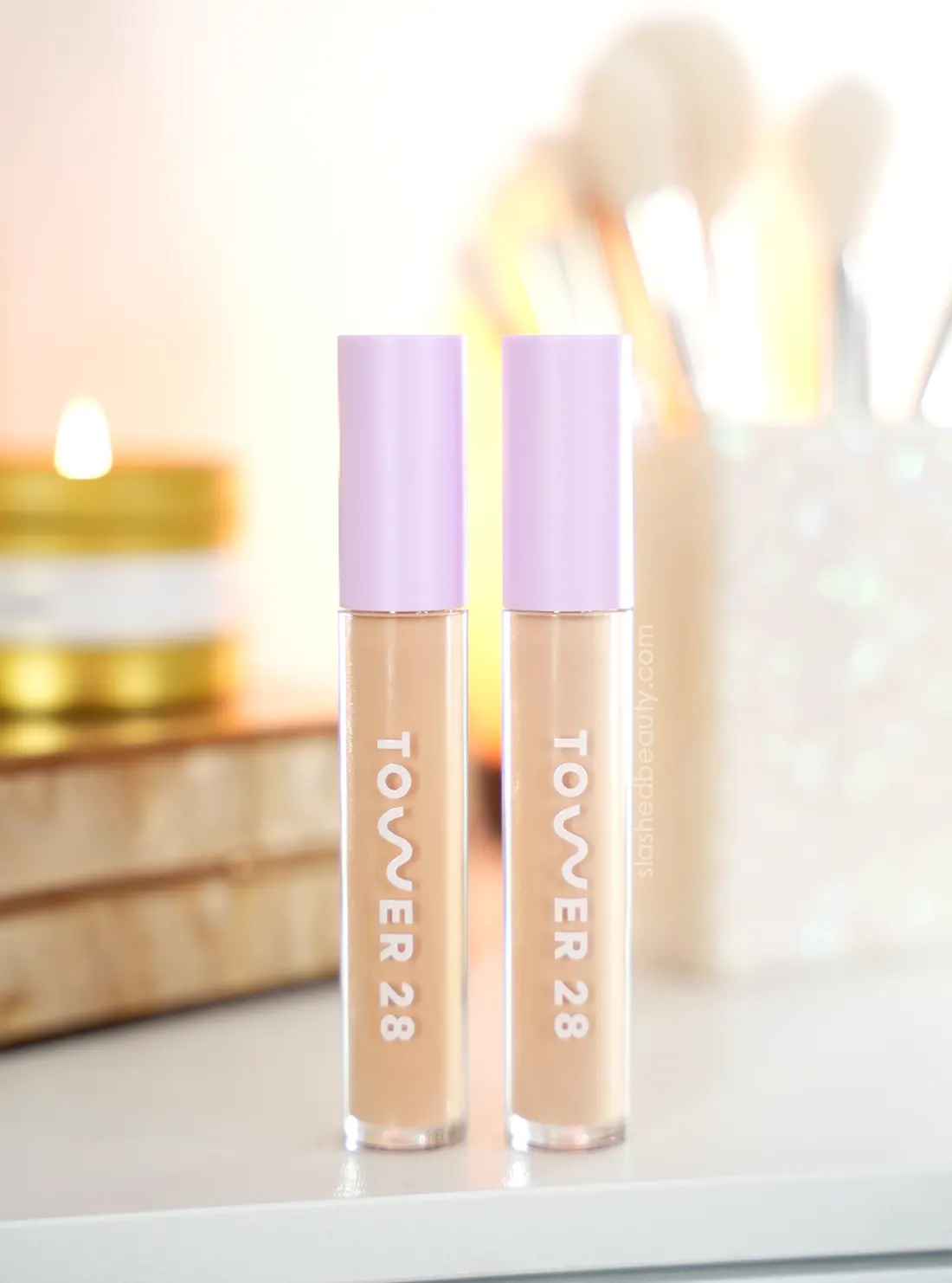 Two tubes of Tower 28 Serum Concealer standing on a vanity with a candle and makeup brushes in the background. | Tower 28 Swipe All Over Serum Concealer Review on Dry Skin | Slashed Beauty 